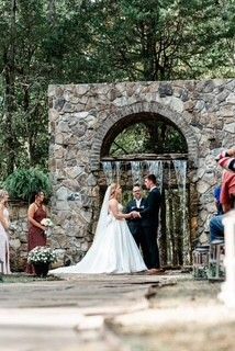 Outdoor wedding ceremony under a stone arch in a forest setting, bride and groom exchanging vows