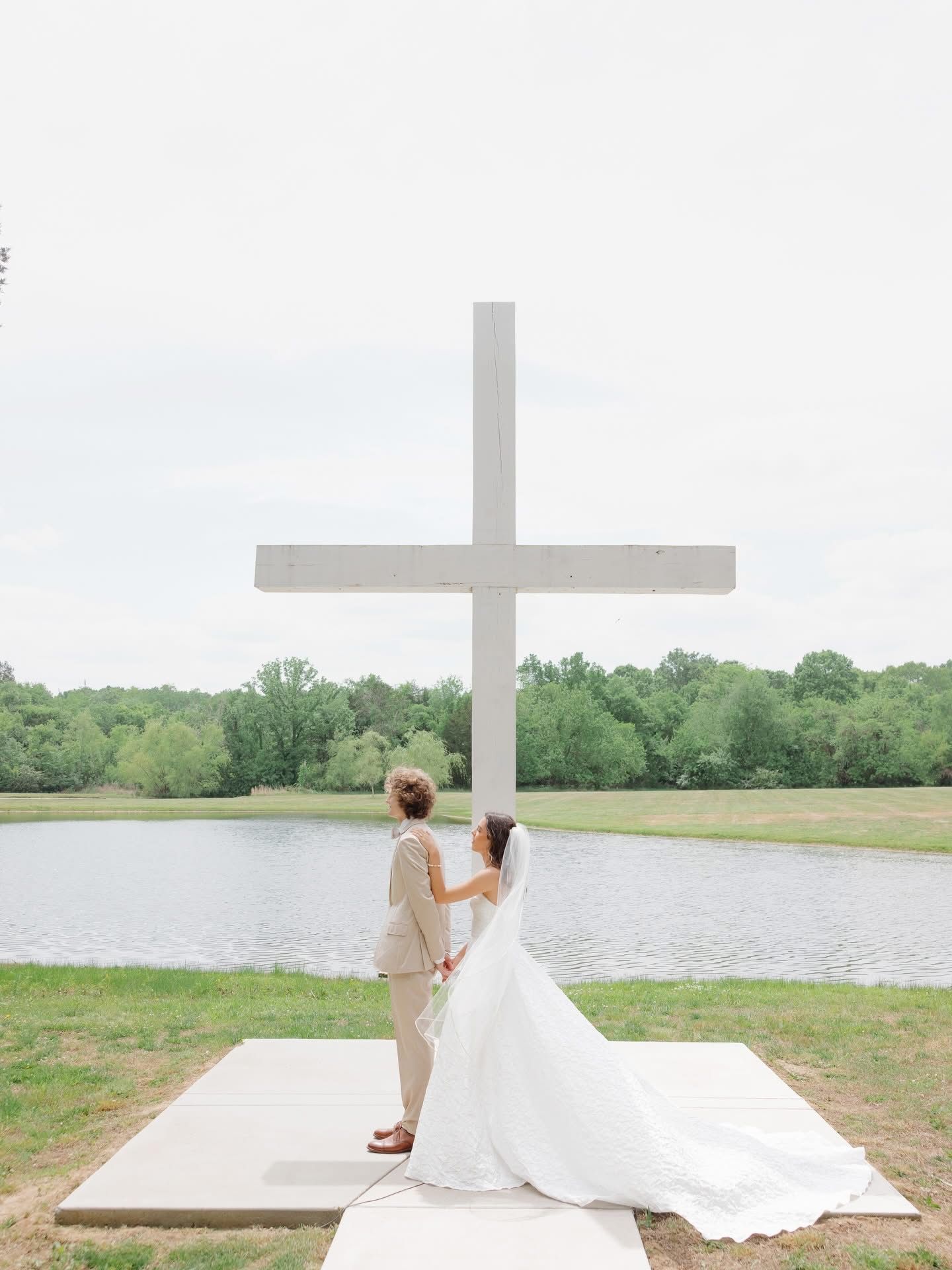 Bride and groom stand before a large cross by a lake, with the bride’s long veil trailing on the ground.