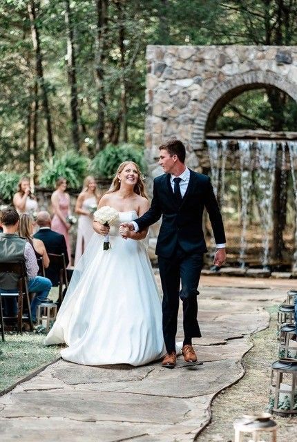 Bride and groom walk down a forest wedding aisle beside a stone archway