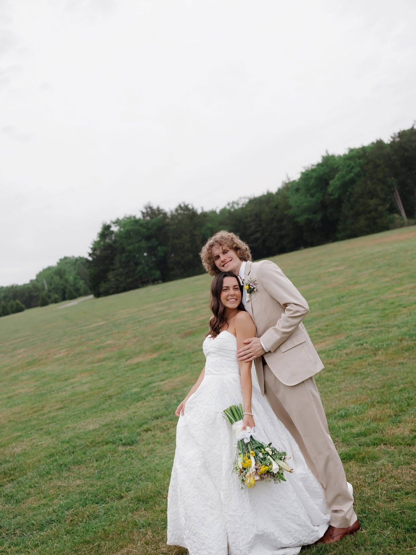 Couple in wedding attire posing in a grassy field, holding a bouquet beneath an overcast sky