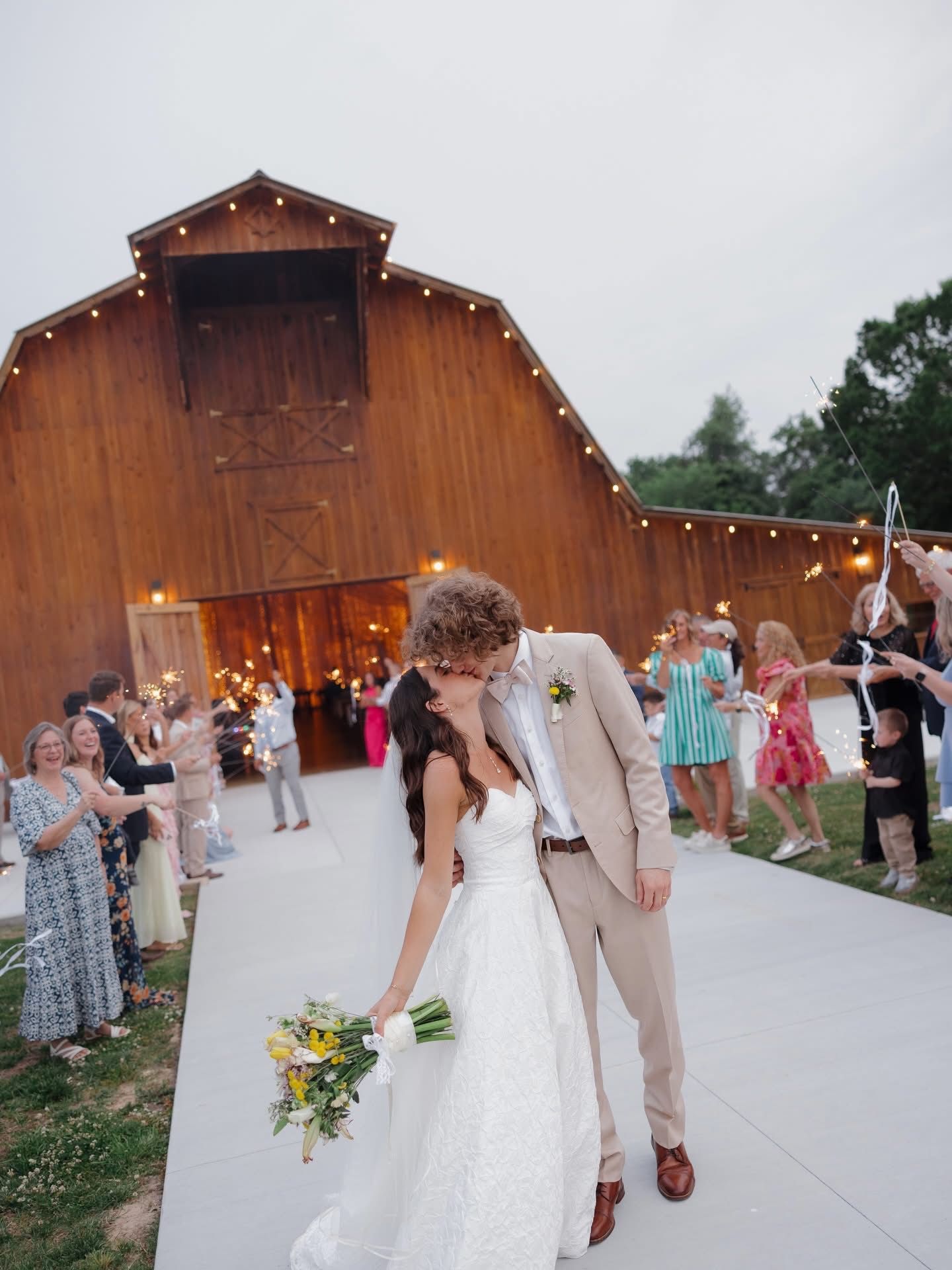 Bride and groom kiss at a rustic barn wedding reception with string lights and guests nearby