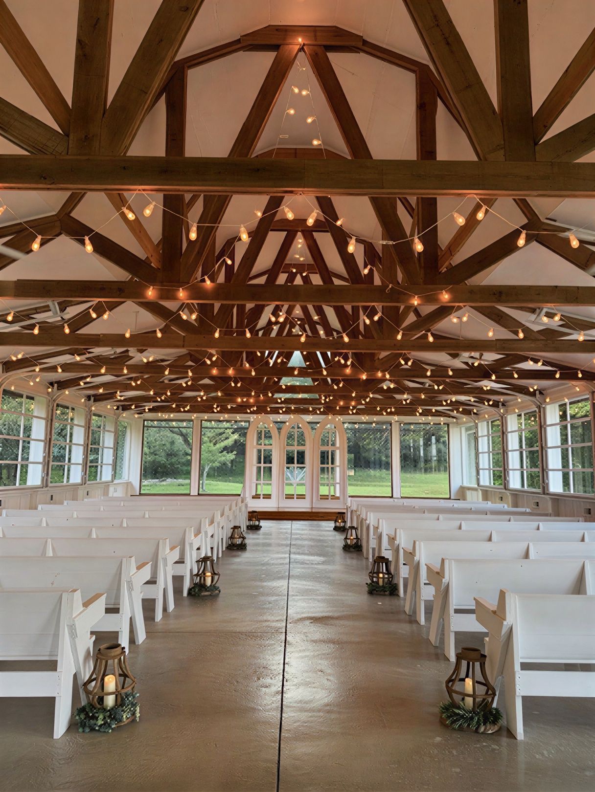 A rustic wedding ceremony hall with white chairs, wooden beams, and string lights, facing arched windows.