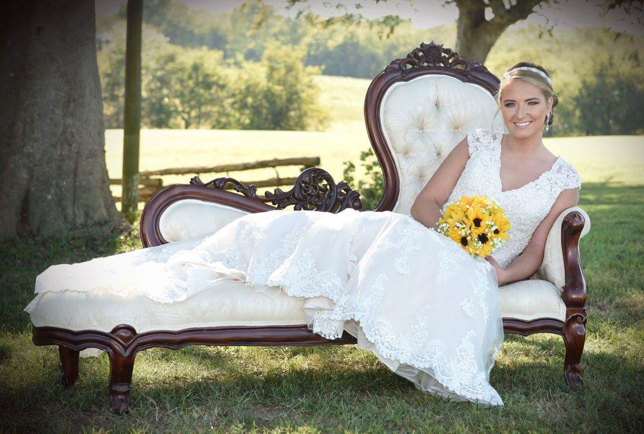 A bride in a wedding dress is sitting on a chaise lounge holding a bouquet of sunflowers.