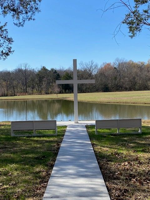 A white cross is in the middle of a grassy field next to a lake.