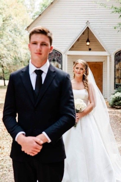 Groom in black tuxedo stands before a bride in white dress and veil outside a church entrance
