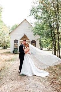 Bride and groom posing outside a small white chapel, her long veil trailing beside them.