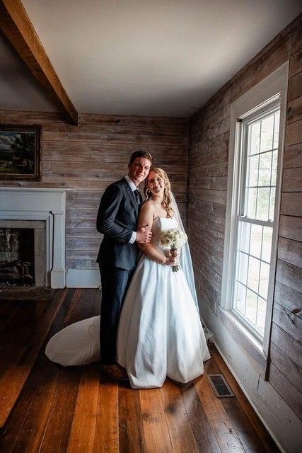 Bride and groom posing together in a room with a fireplace and tall window, holding a bouquet