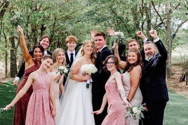 Wedding party posing outdoors among trees, bride in white and bridesmaids in pink, smiling and celebrating