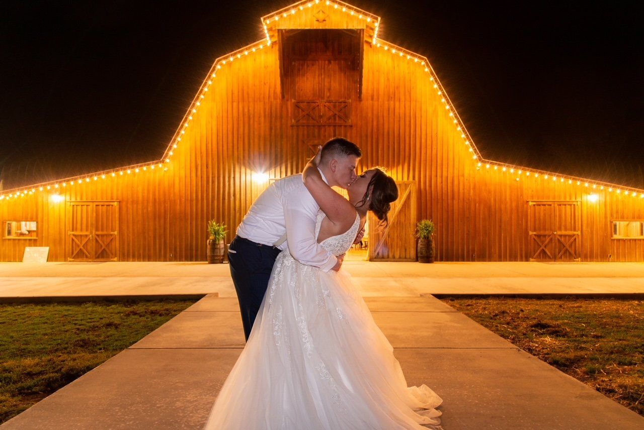 A bride and groom are kissing in front of a barn lit up at night.