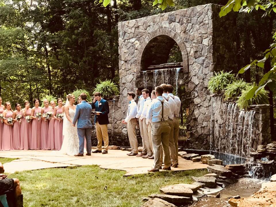 A bride and groom are standing in front of a waterfall during their wedding ceremony.