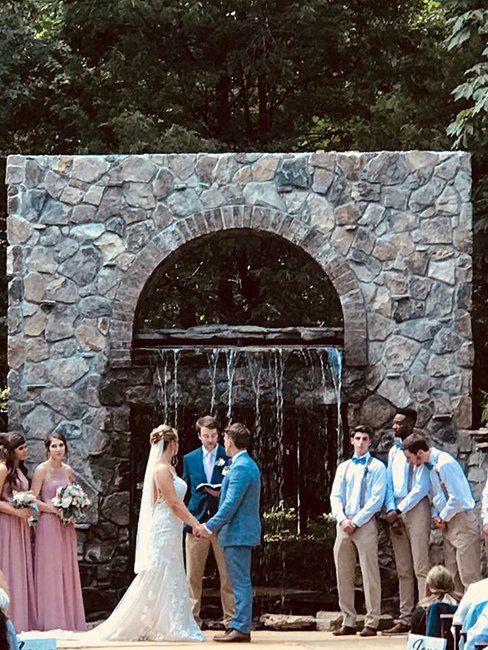 A bride and groom are holding hands during their wedding ceremony in front of a waterfall.