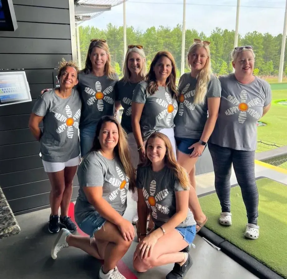Group of women in matching daisy shirts at a golf driving range; smiling, posing for photo.