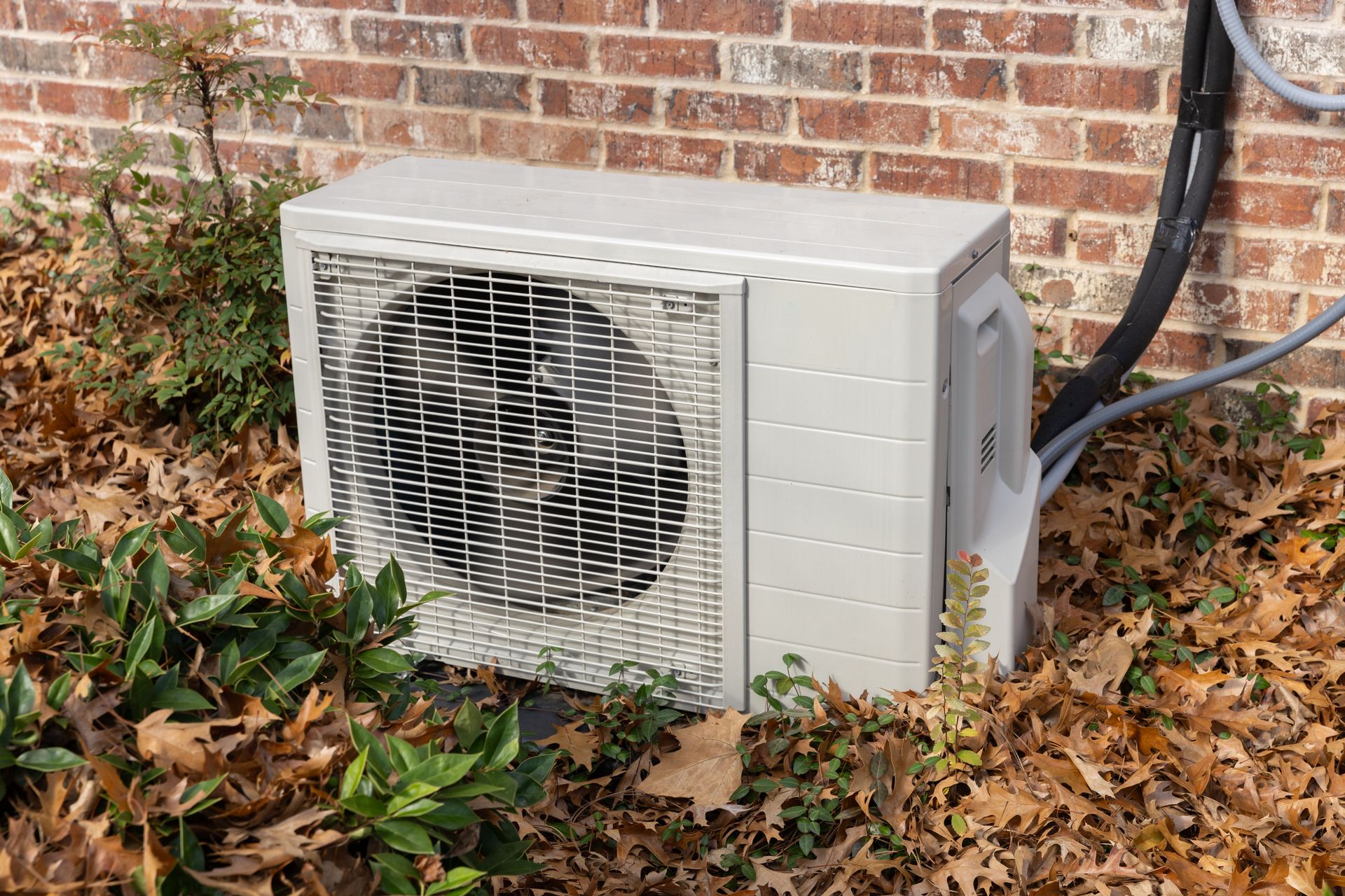 Outdoor air conditioner unit beside a brick wall, surrounded by fallen leaves and shrubs