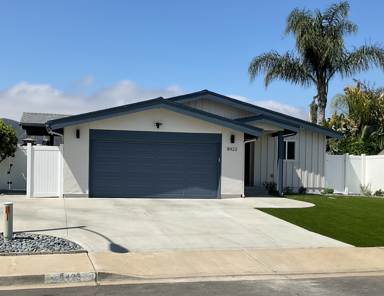 Single-story suburban house with a gray garage door, white exterior, driveway, and palm trees in front