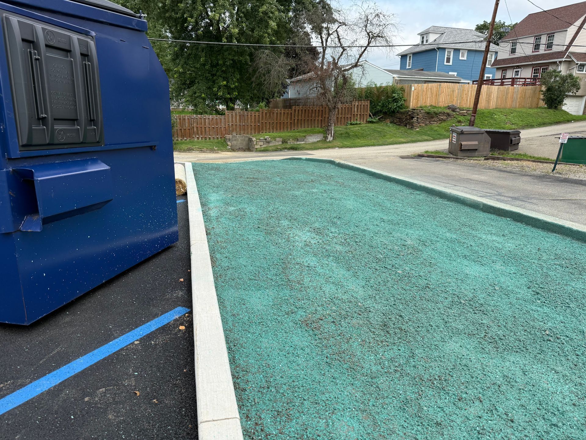 Blue dumpster beside a teal gravel lot near a white curb and parking area outdoors