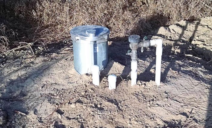 A metal bucket is sitting in the dirt next to a pipe.