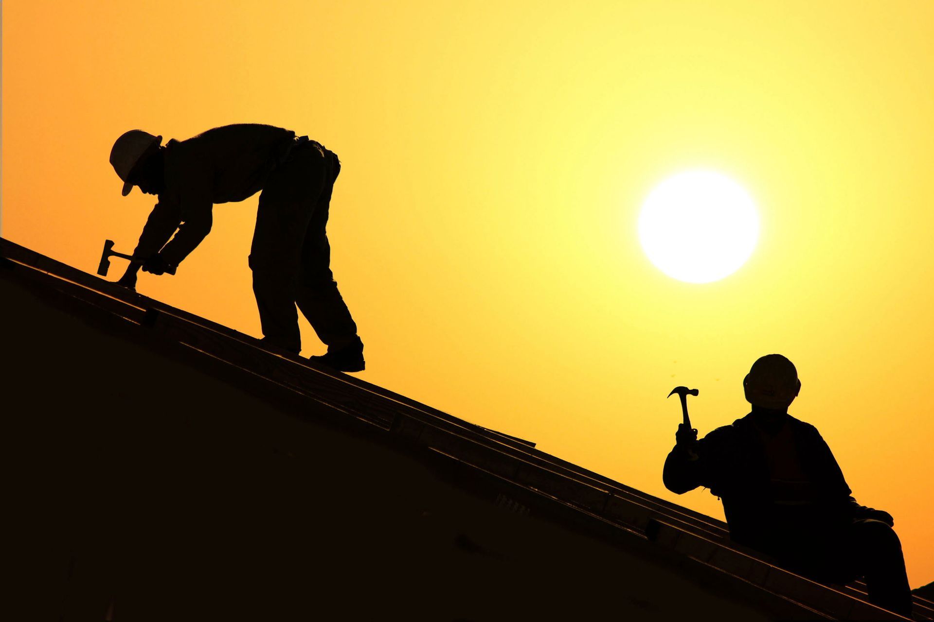 Silhouette of two construction workers on a roof holding hammers at sunset with the sun in the background.
