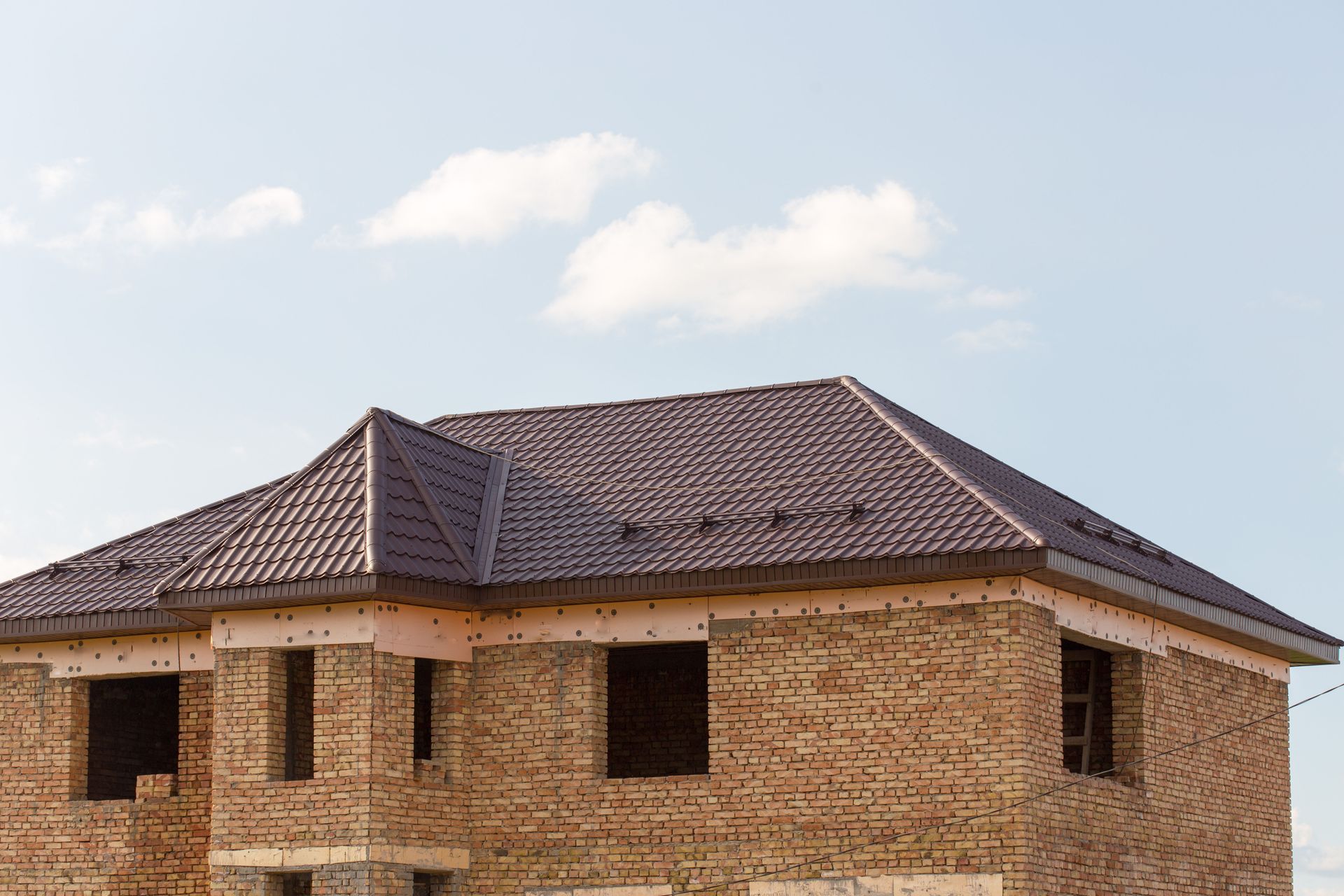 Brick house under construction with a new dark brown metal tile roof against a blue sky.