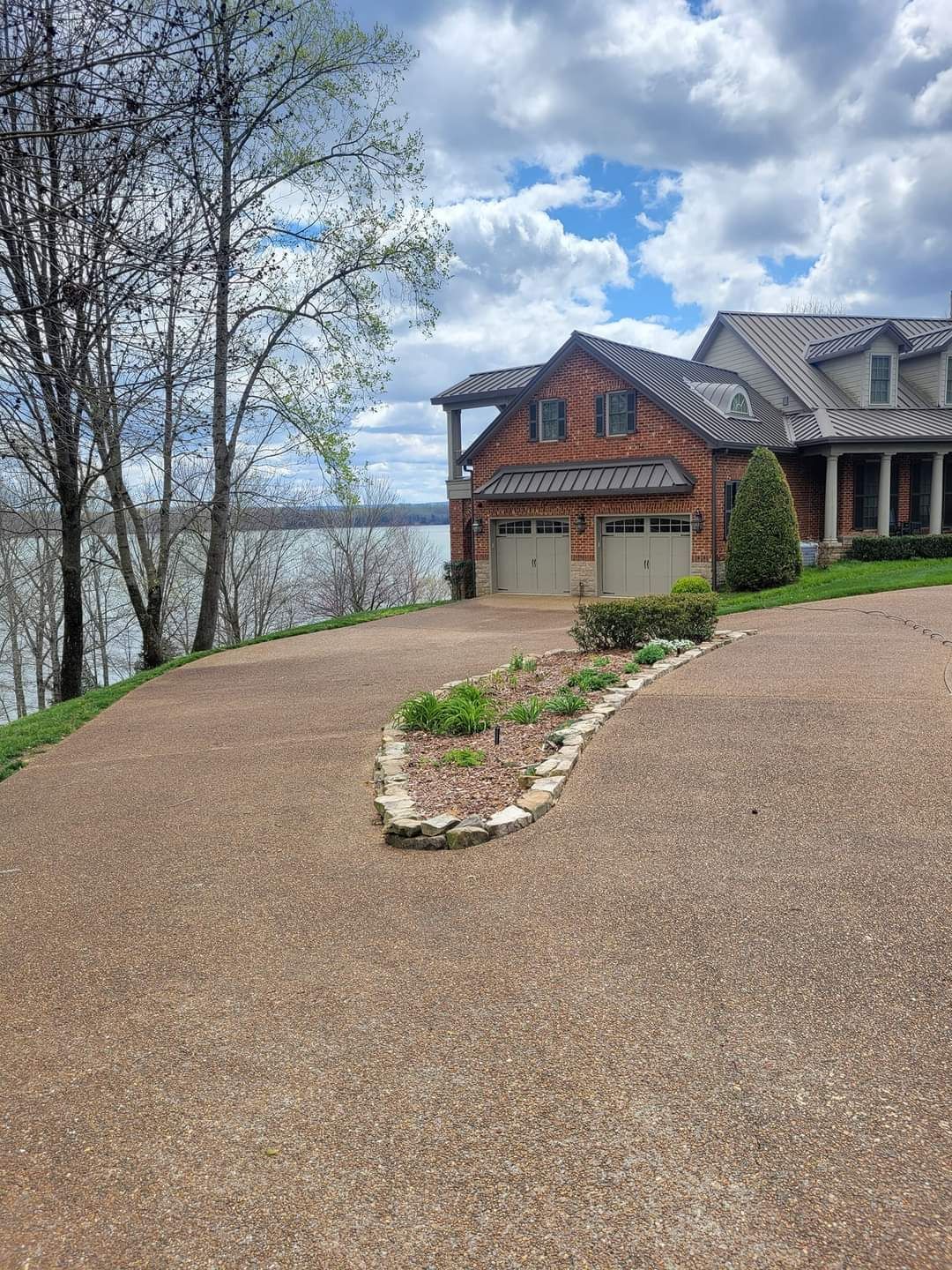Large stone driveway leading to a lakeside home with trees and cloudy sky beyond