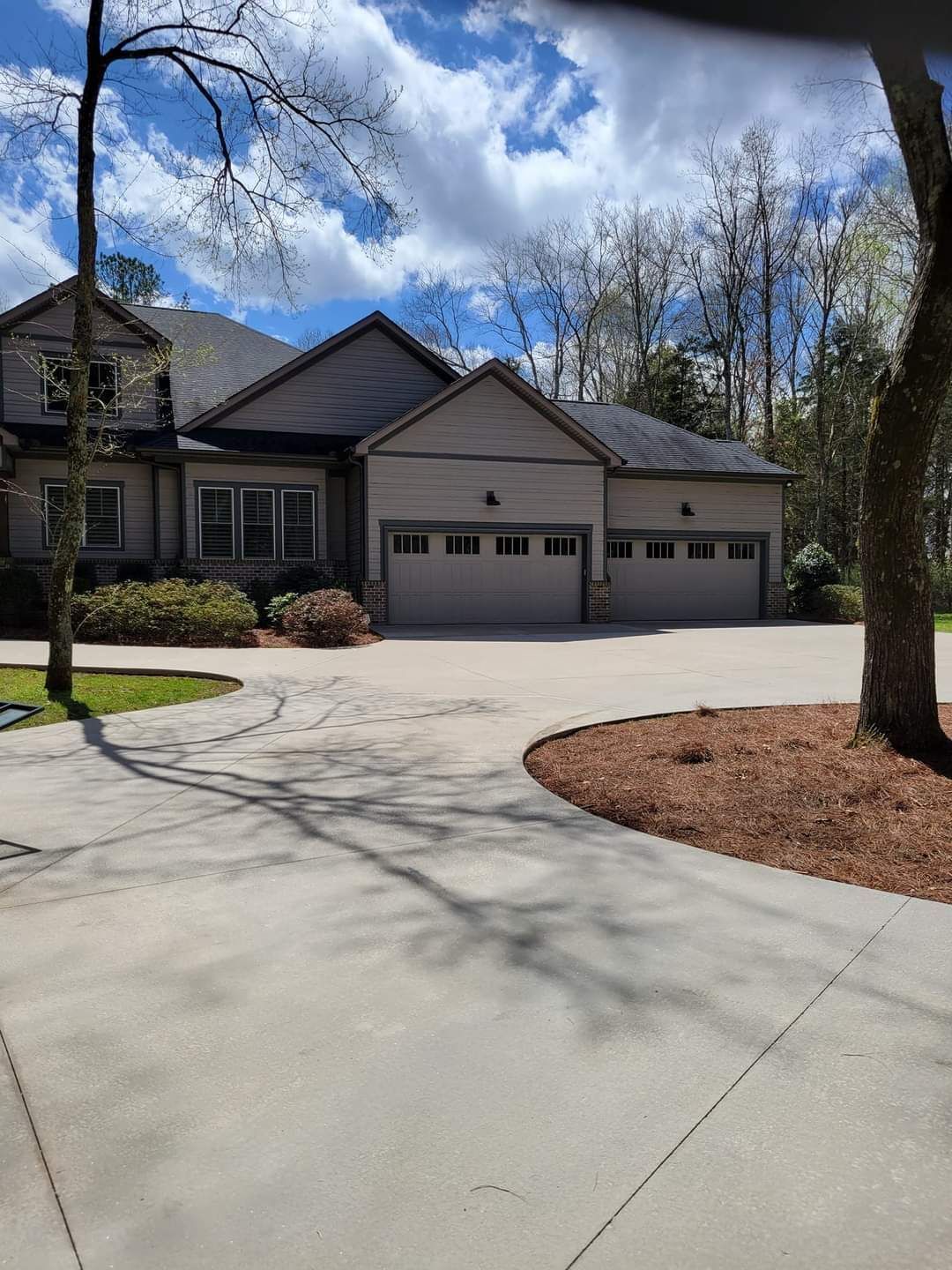 Suburban house with two-car garage, curved driveway, and trees under a cloudy blue sky