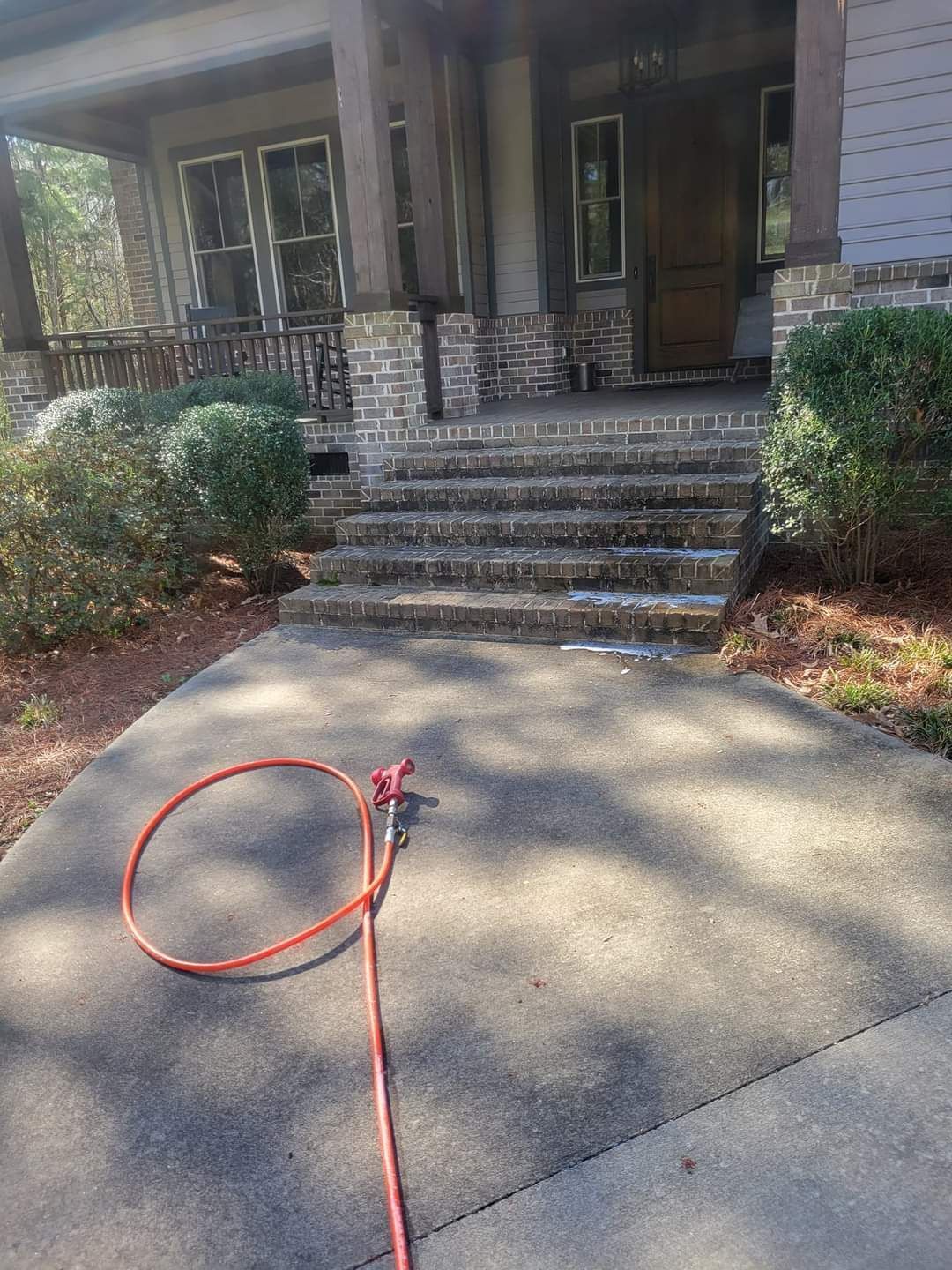 Red hose on a concrete driveway leading to a house porch with steps and shrubs.