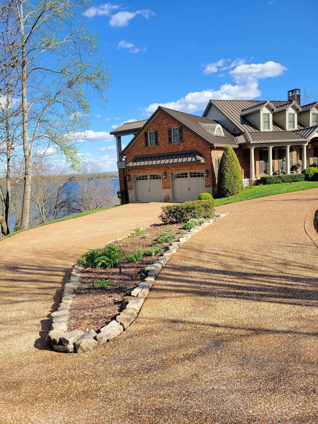 Large suburban house with stone driveway, garage, and hillside lake view on a sunny day