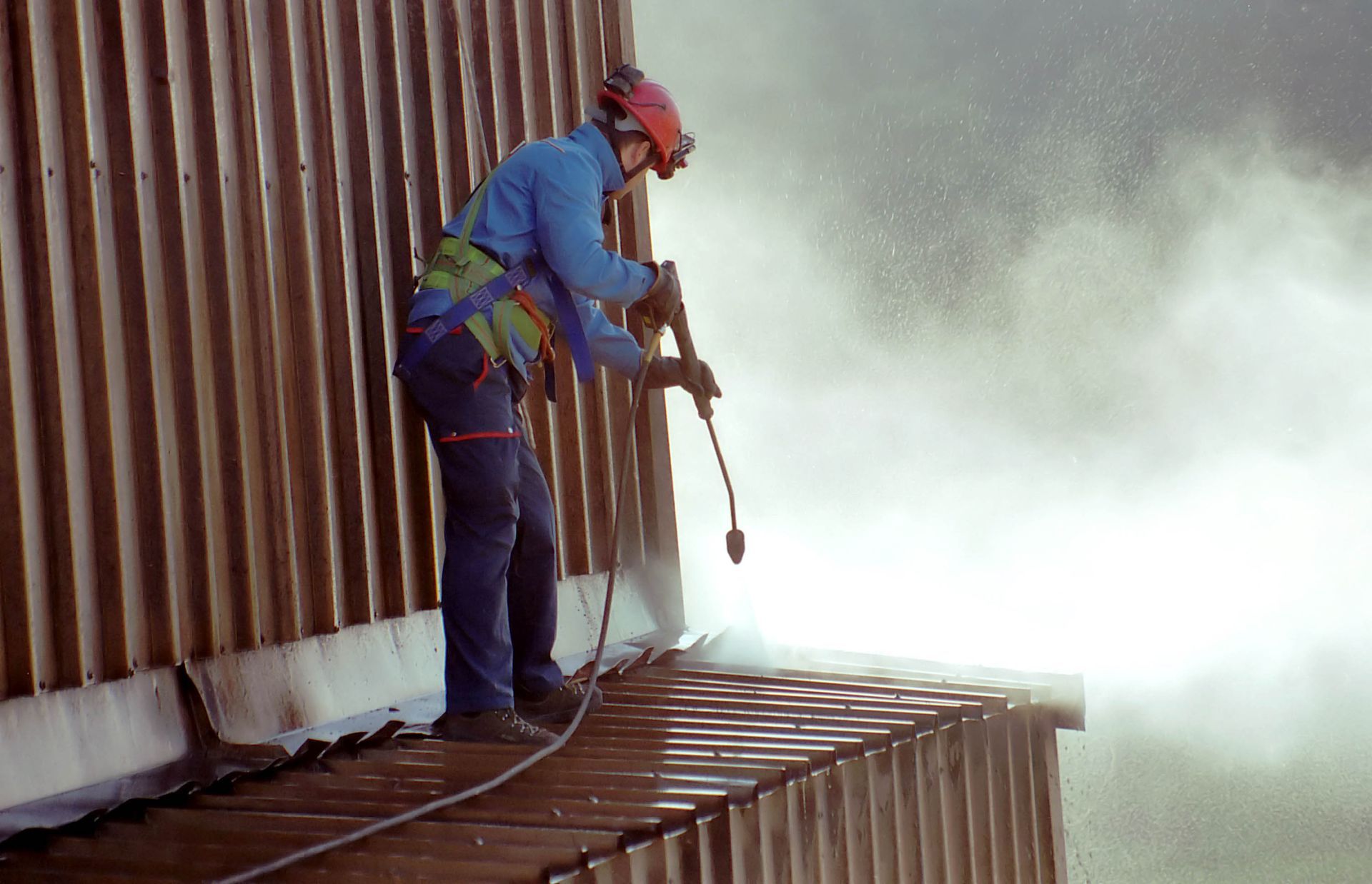 Worker in safety gear spray-paints a metal container on a platform, with mist in the background.