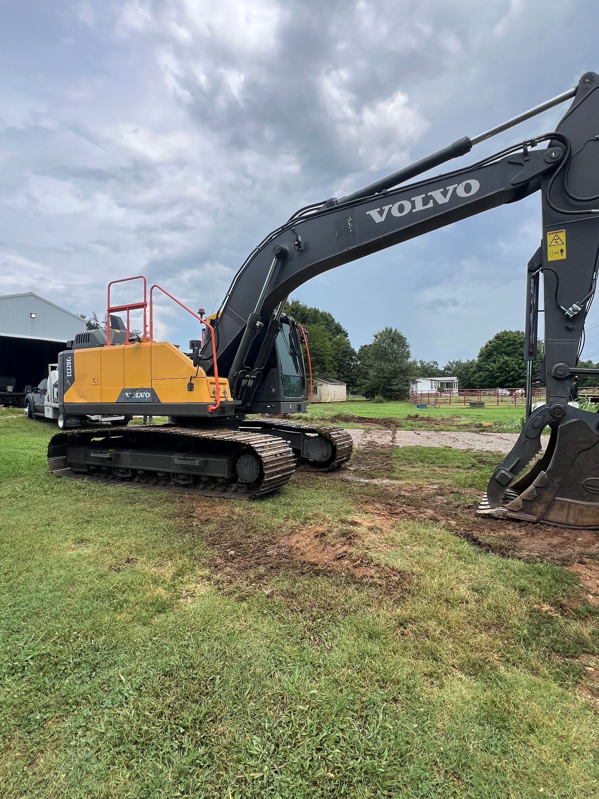 Yellow Volvo excavator on tracks digging a muddy area beside grass under a cloudy sky