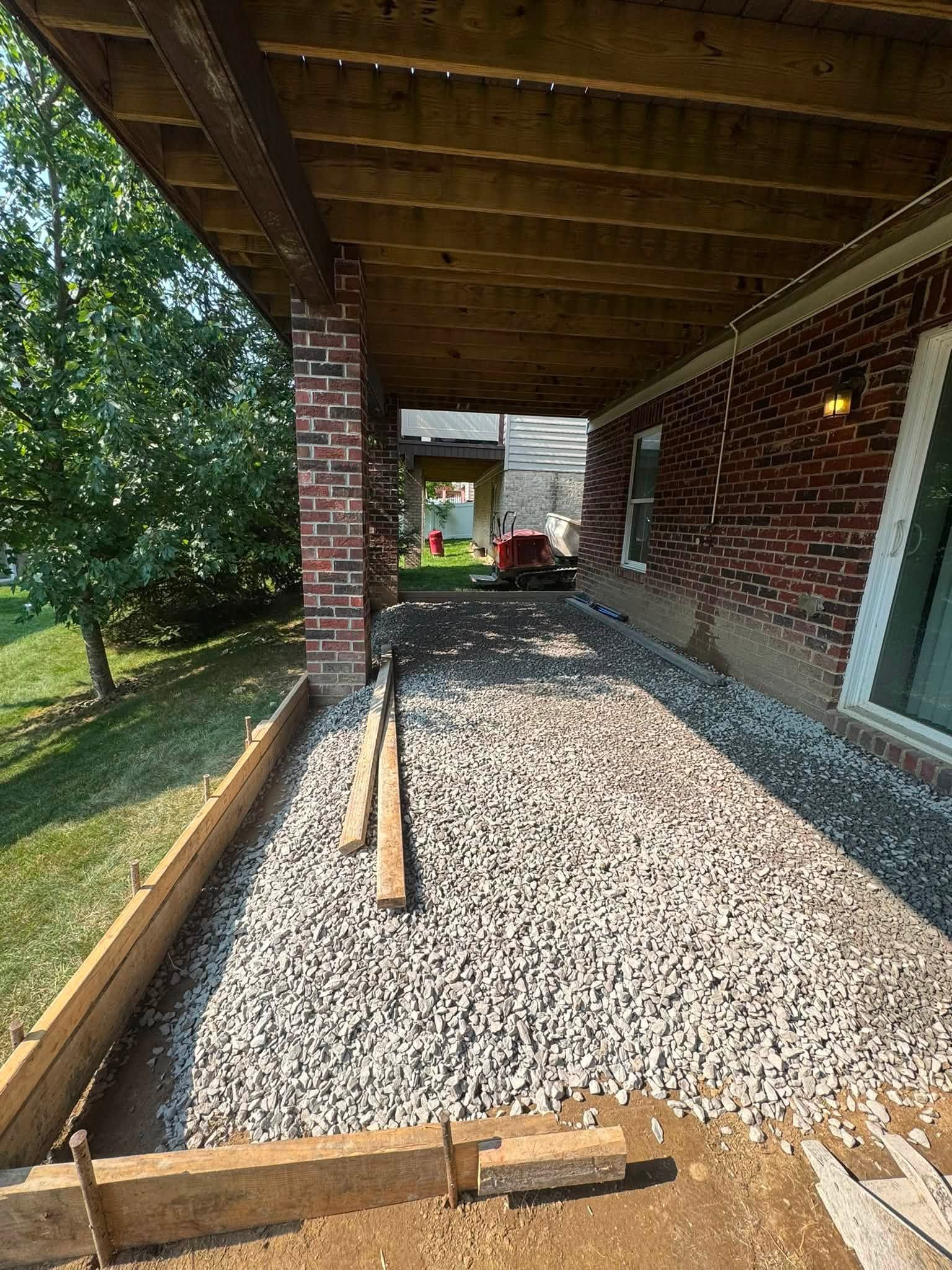 Covered brick patio with gravel walkway, wooden edging, and trees beside a house