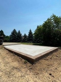 Concrete slab foundation in a dirt lot, surrounded by trees under a clear blue sky