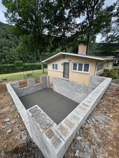 Small house beside a gray concrete foundation pit with stone retaining walls in a wooded area