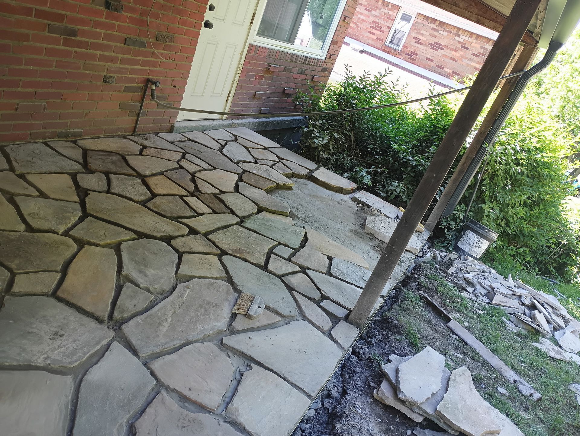 Stone patio beside a brick house, with a small garden and wooden posts along the edge.