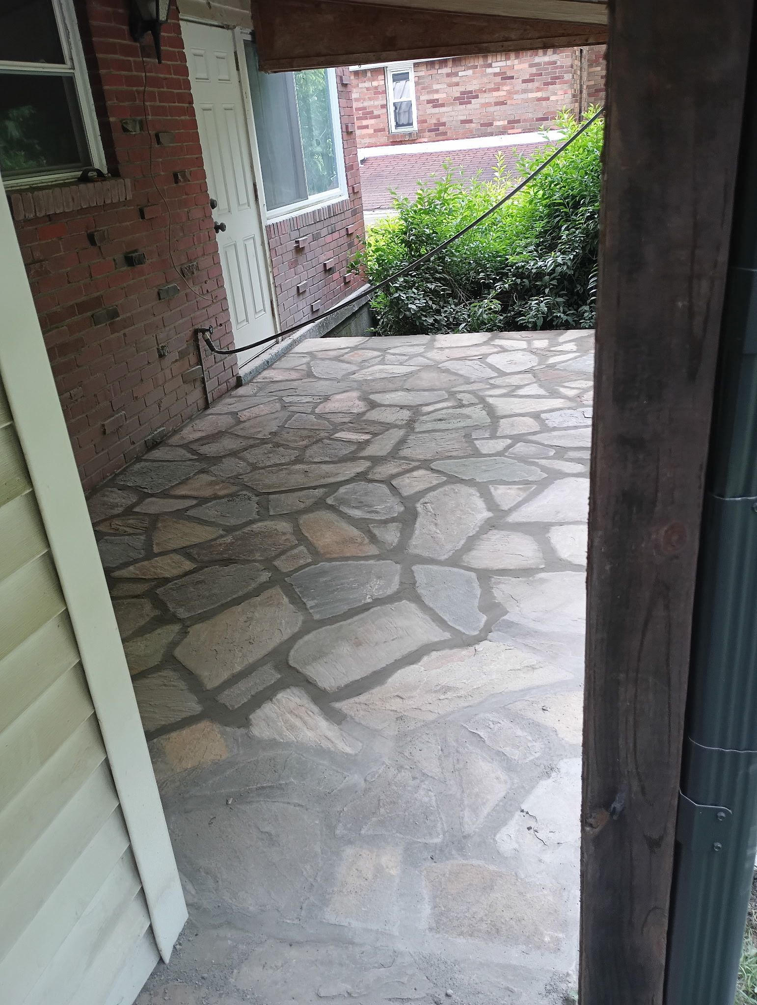 Stone patio walkway beside brick house, framed by a porch opening, with green shrubs ahead.