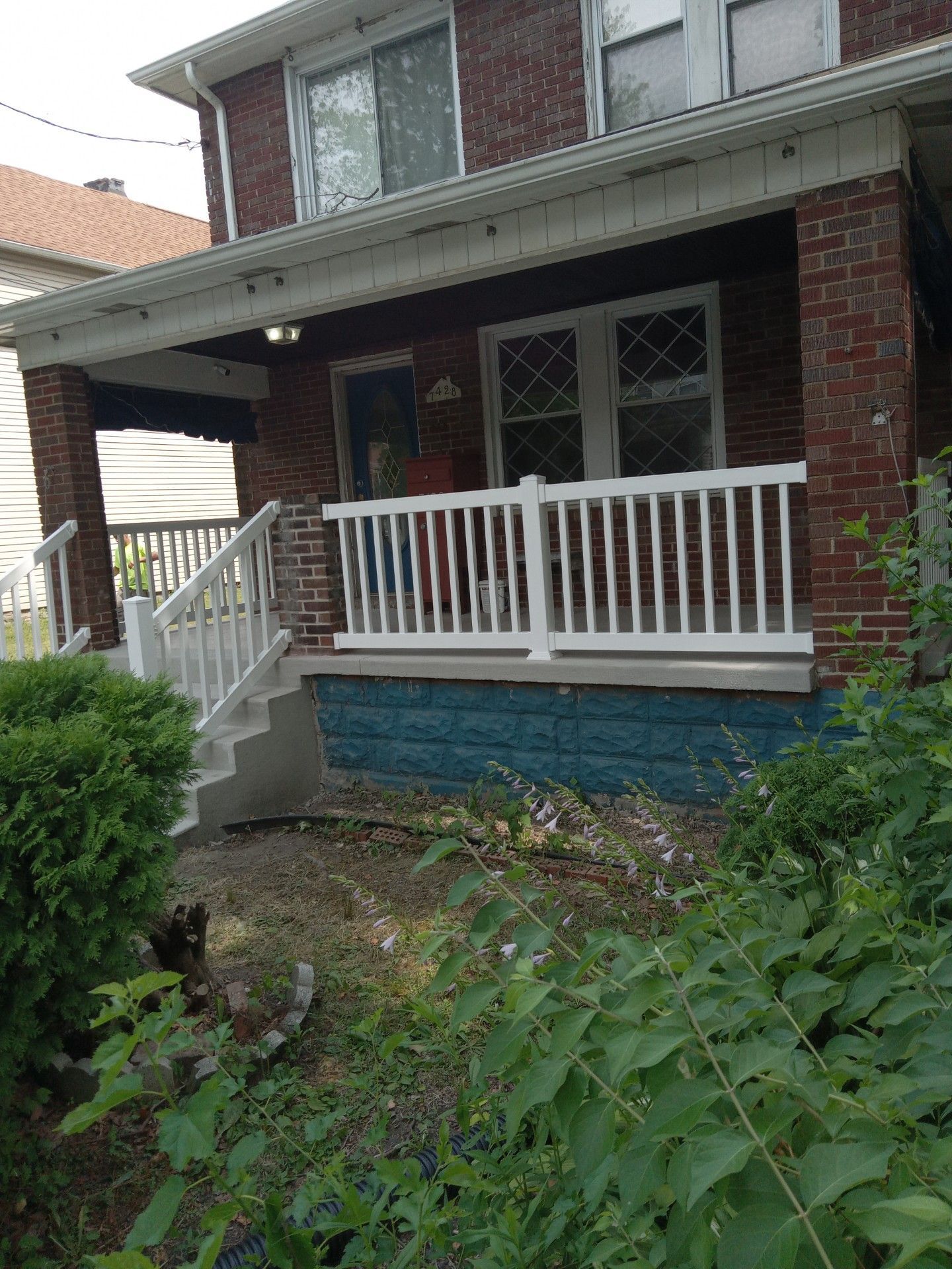 Brick house porch with white railing, blue foundation, and overgrown front garden