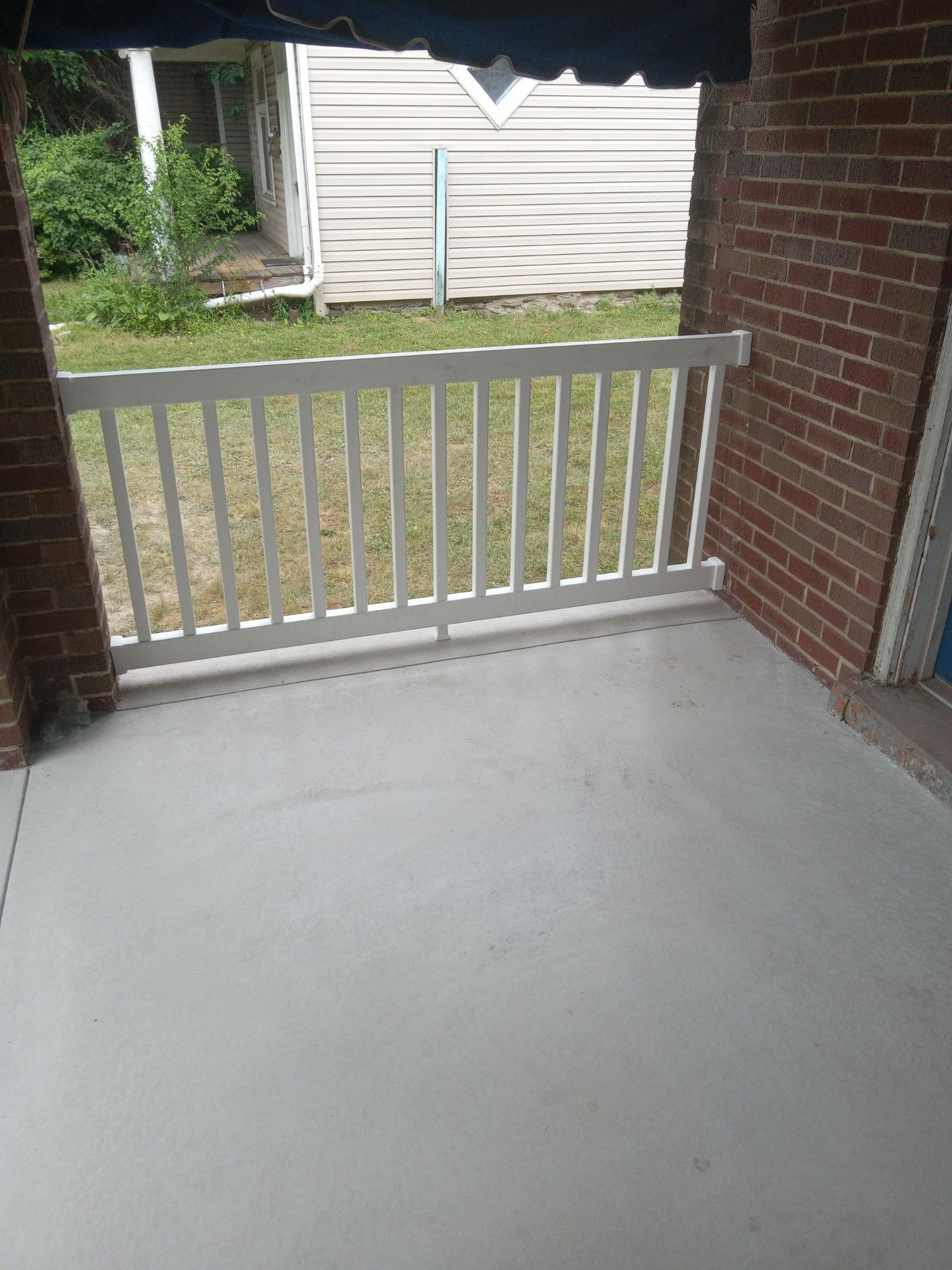Porch with white railing and concrete floor, overlooking a grassy yard and white shed beside a brick wall