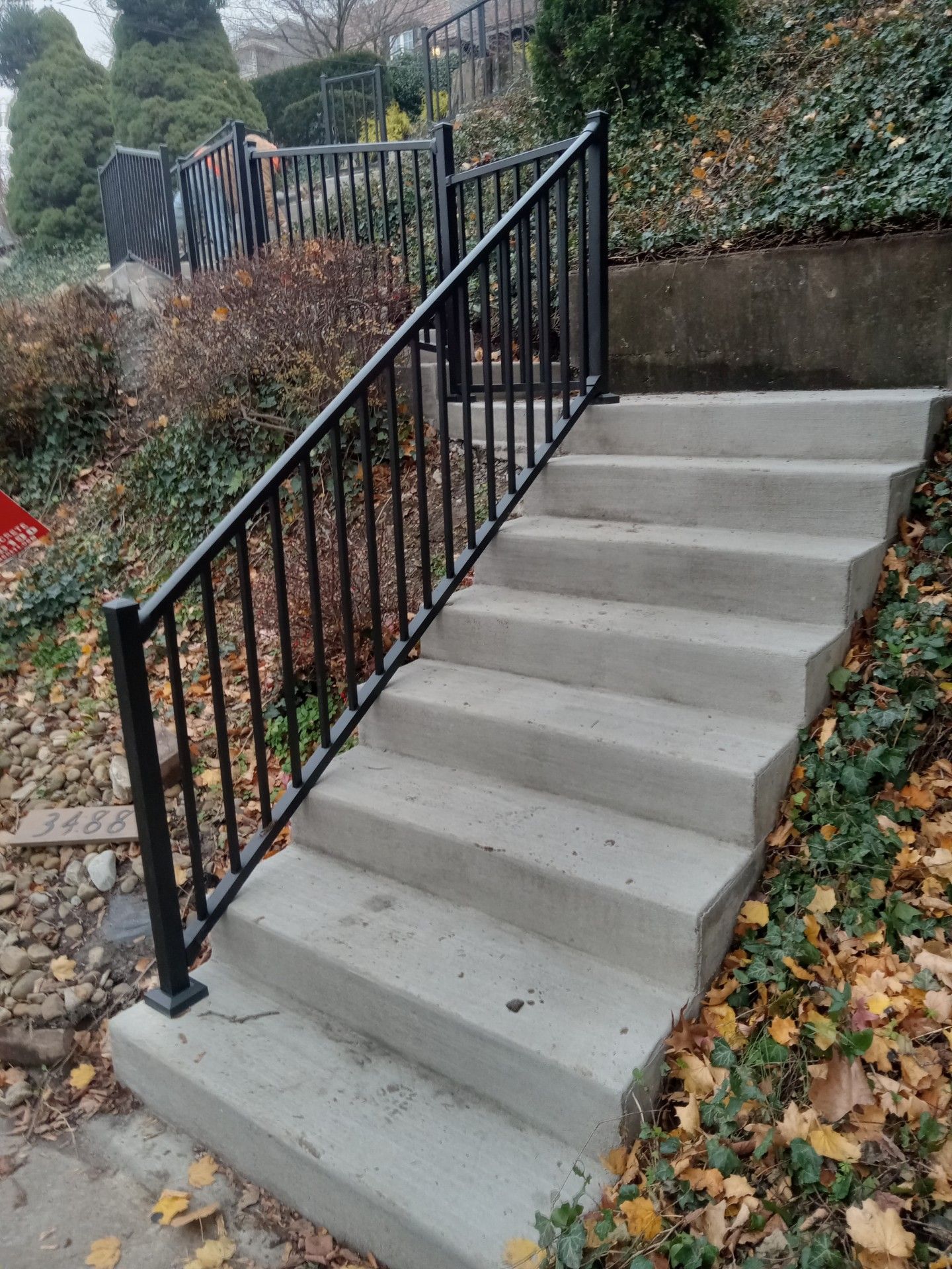 Concrete outdoor stairs with black metal railing beside a garden path covered in fallen leaves