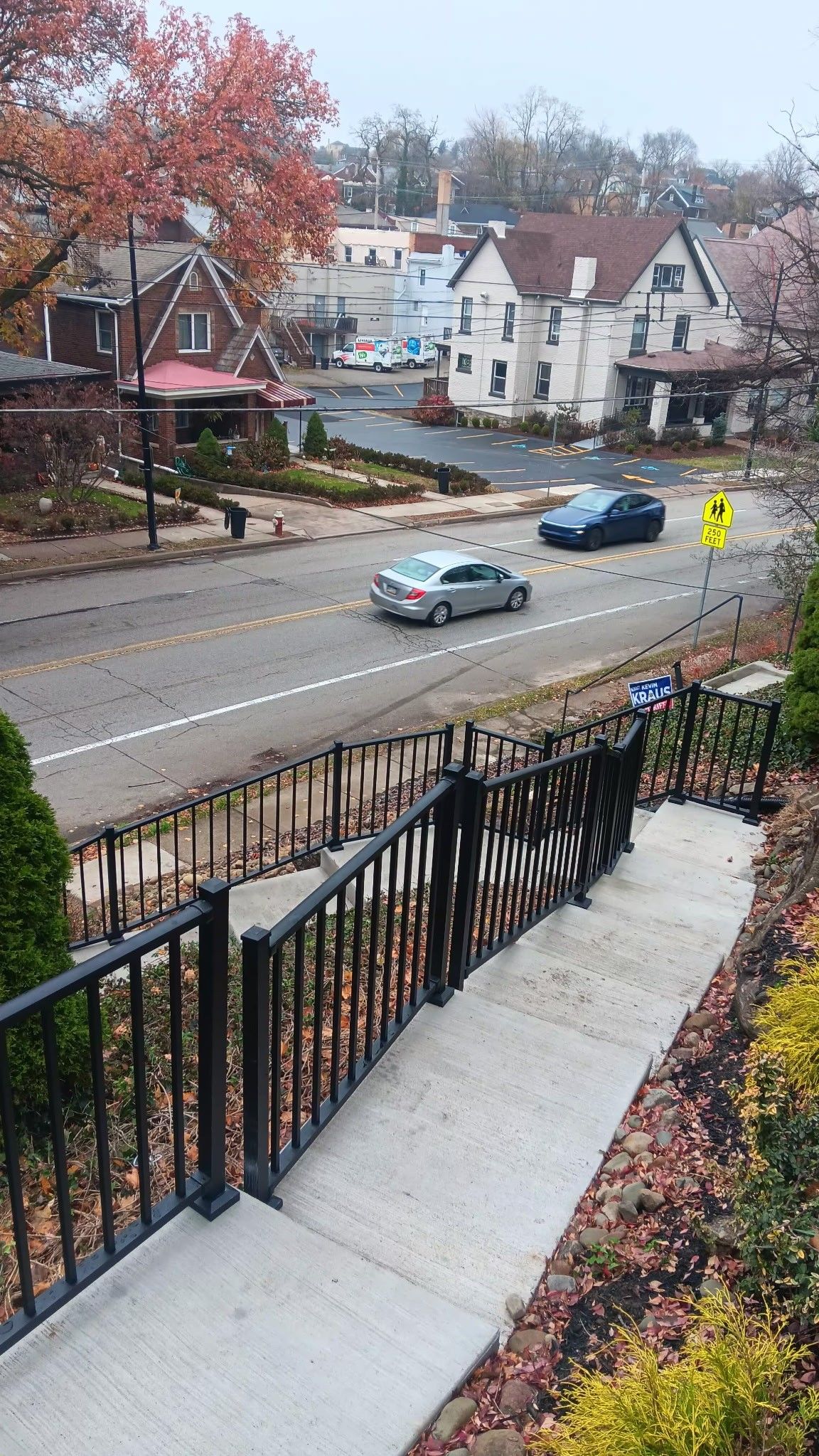 Residential street with two cars passing by, viewed from a front porch with steps and autumn trees.