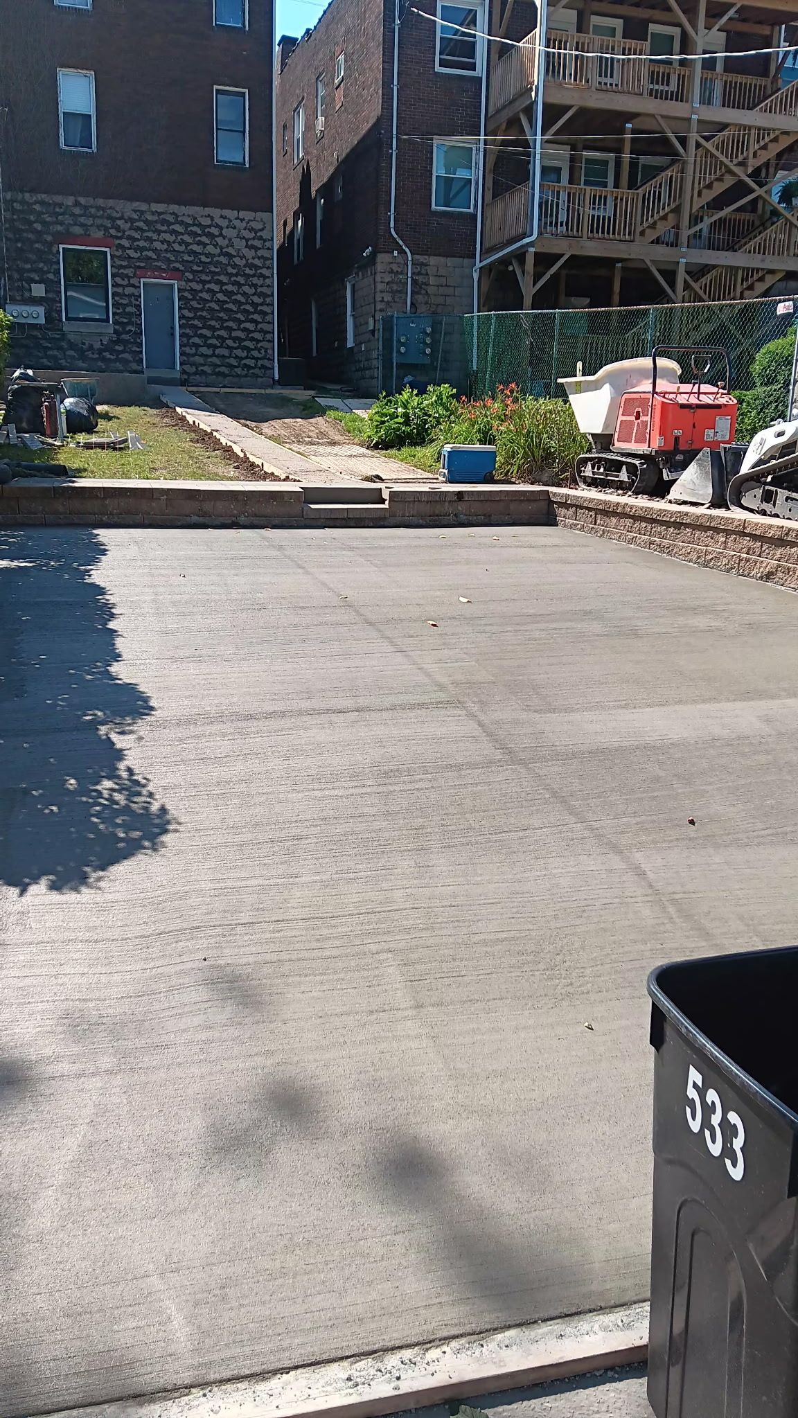 Construction site courtyard with gravel path, brick buildings, scaffolding, and equipment in bright sun