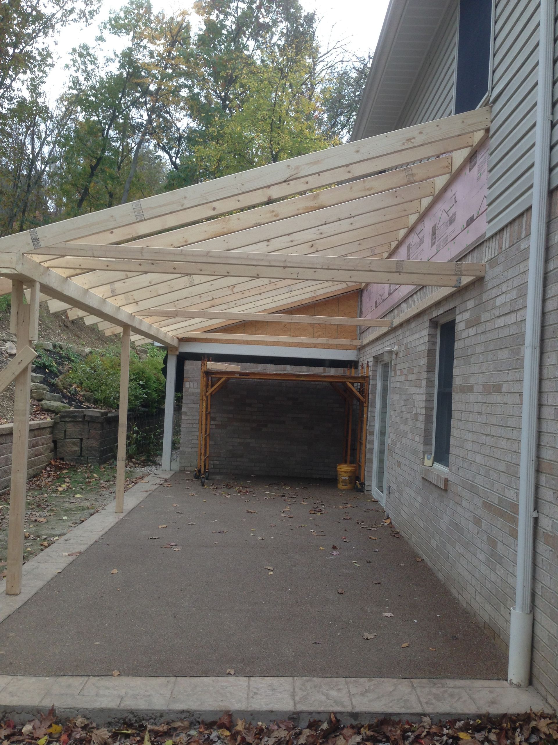 Covered carport with slanted wood roof framing beside a brick house, over a gravel driveway.