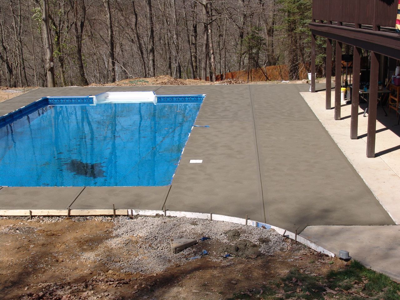 Outdoor pool with blue water beside a concrete deck and covered patio in a wooded area