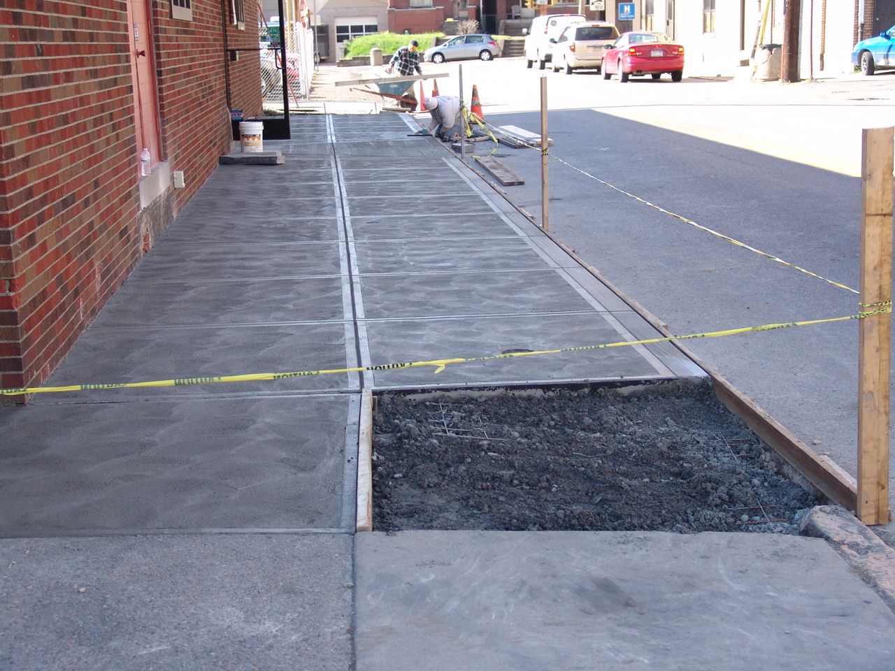 Sidewalk under construction beside a brick building, with taped-off sections and gravel patches.