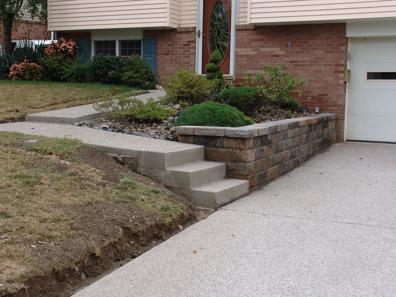 Front yard landscaping with stone retaining wall, concrete steps, and driveway beside a brick house.