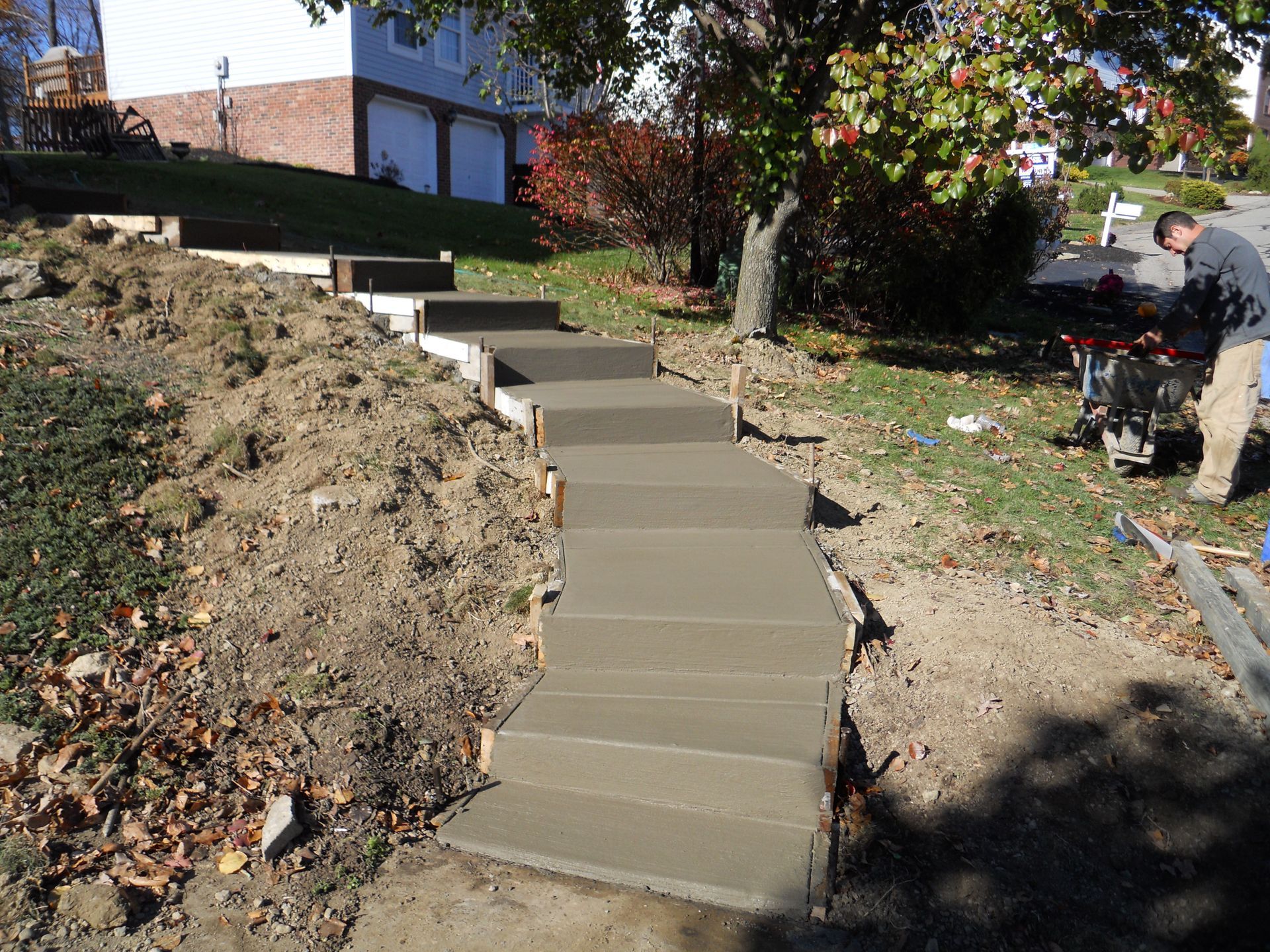 Concrete steps built into a dirt slope beside a house, with a tree and garden tools nearby.