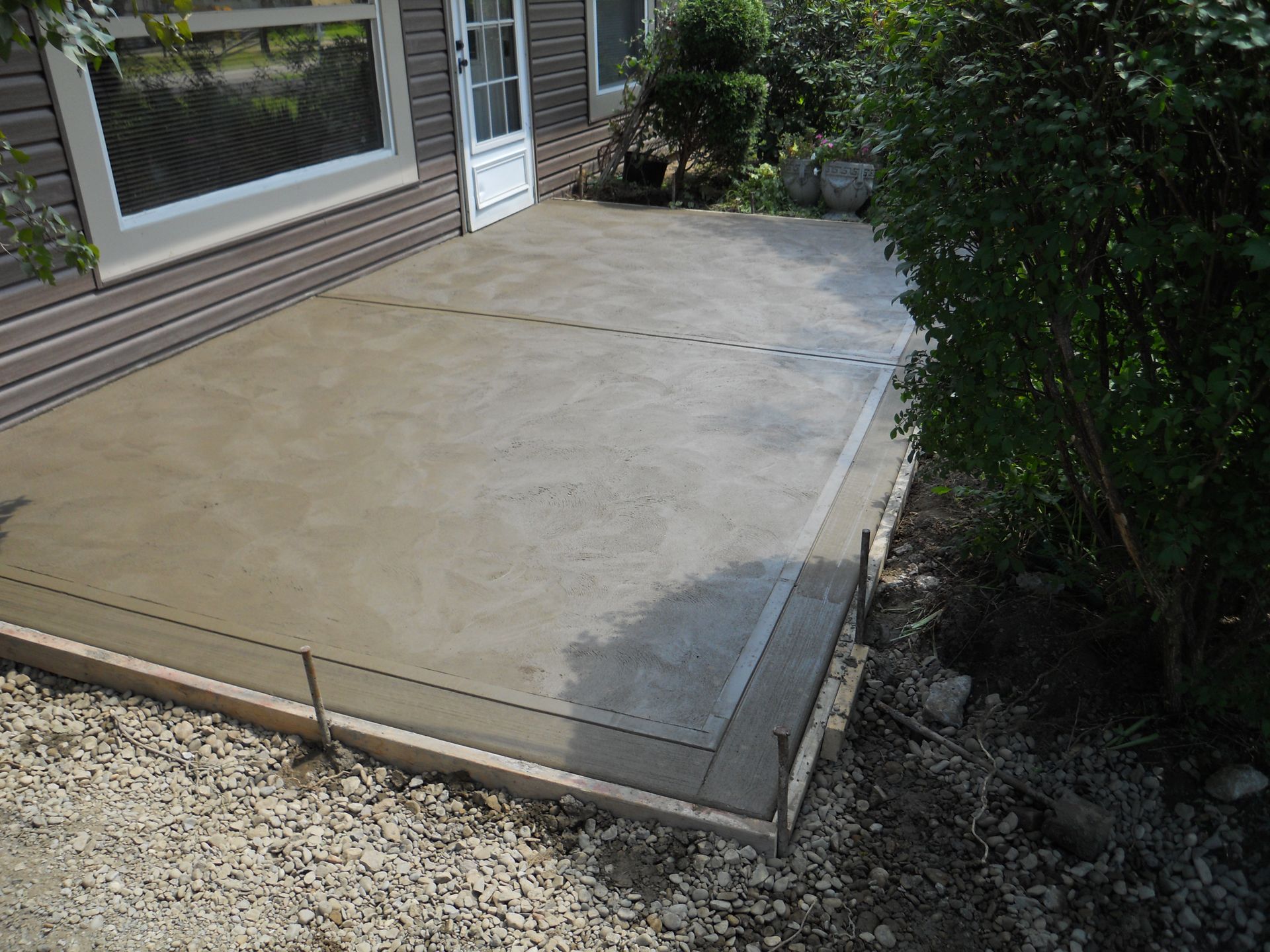 Concrete patio beside a house with a gravel border and shrubs