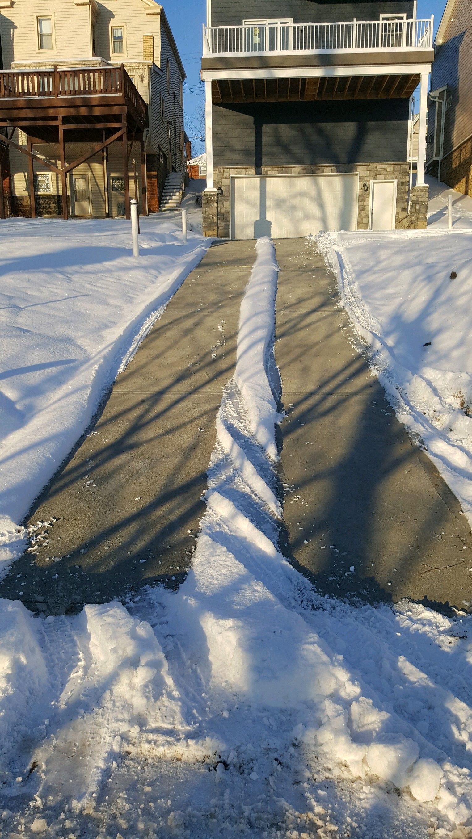 Snowy driveway with tire tracks leading to a house and porch in bright sunlight.