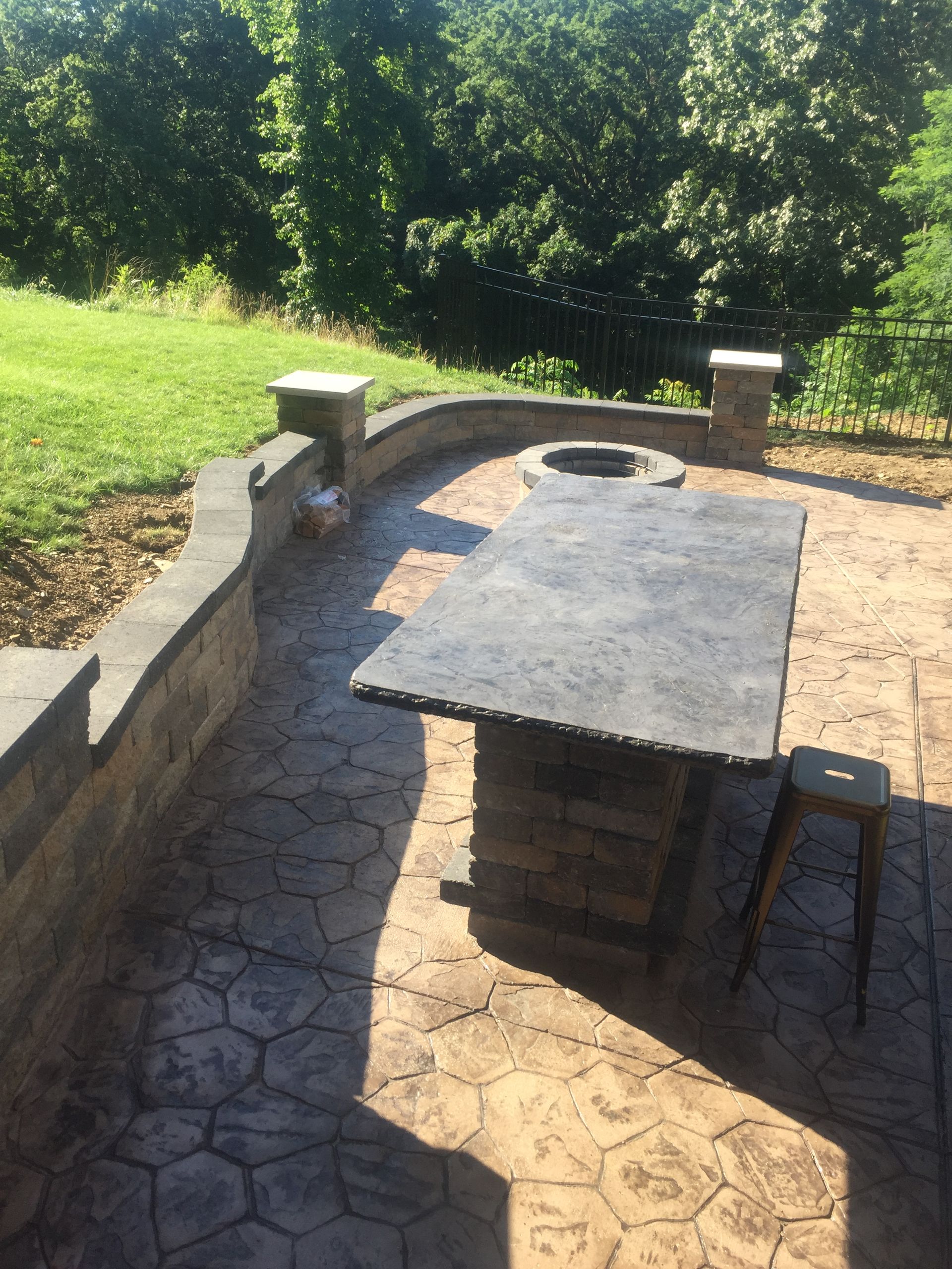 Stone patio with a brick table and stools overlooking a green wooded area in sunlight