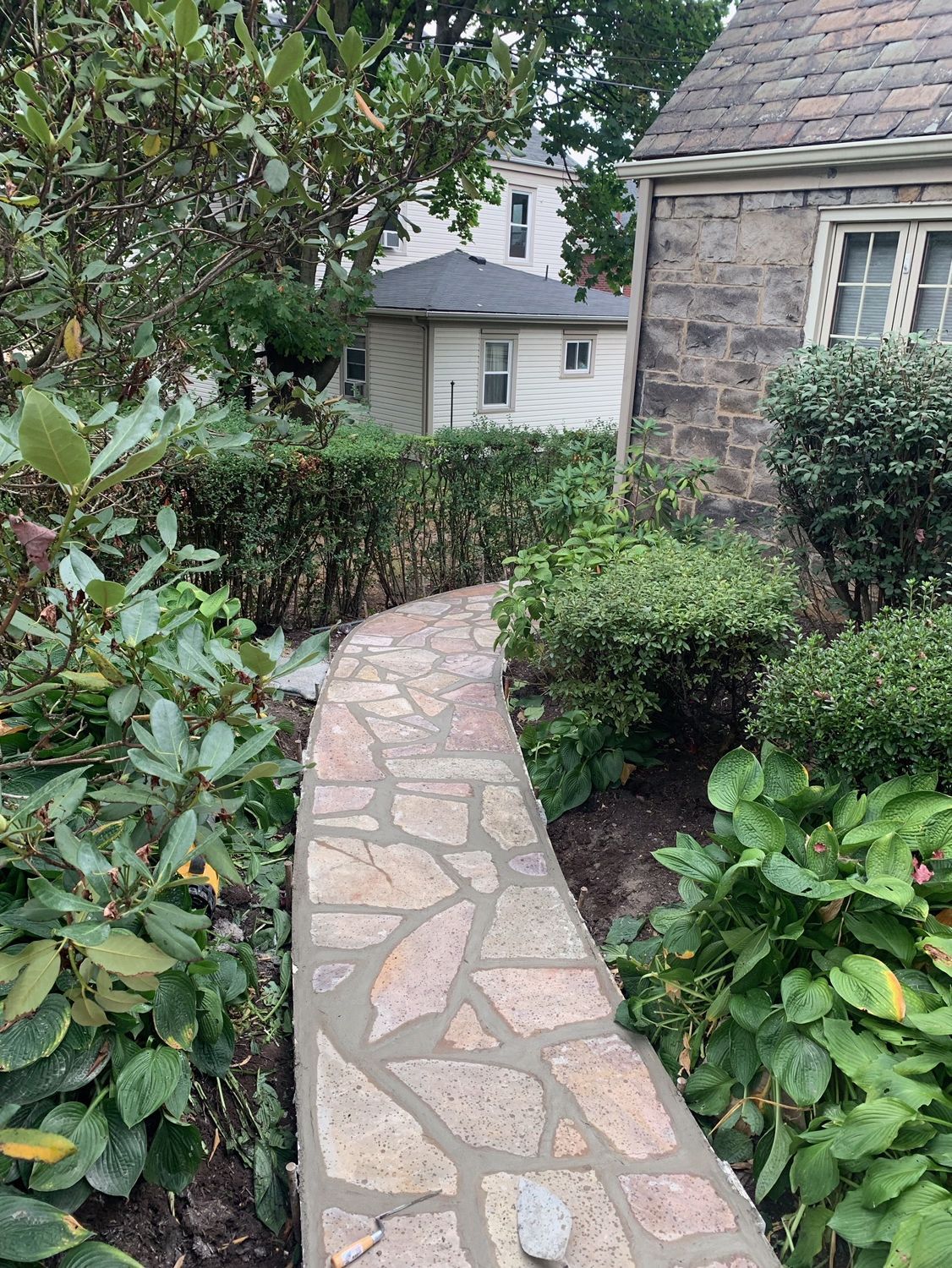 Curved stone walkway through a green garden beside a stone house and shrubs