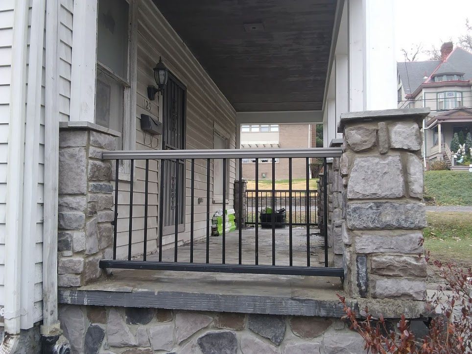 Stone porch with black metal railing beside a house entrance, with steps and a small yard.