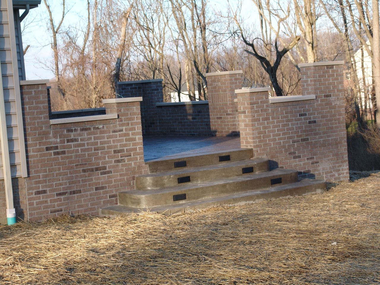 Brick steps leading to a raised patio beside a brick wall, with leafless trees in the background.