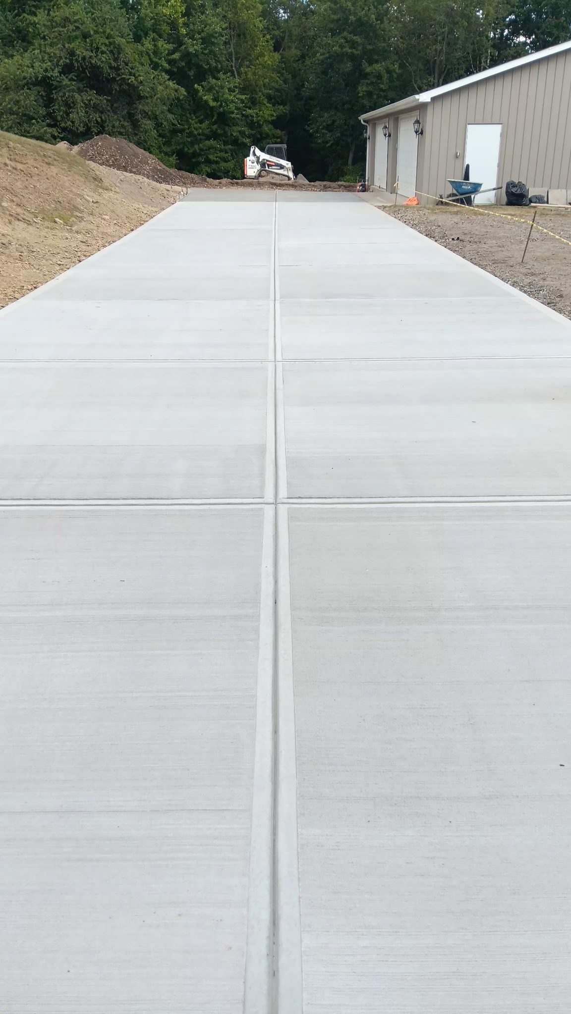 Concrete driveway leading uphill beside a building and dirt slopes, with trees in the background